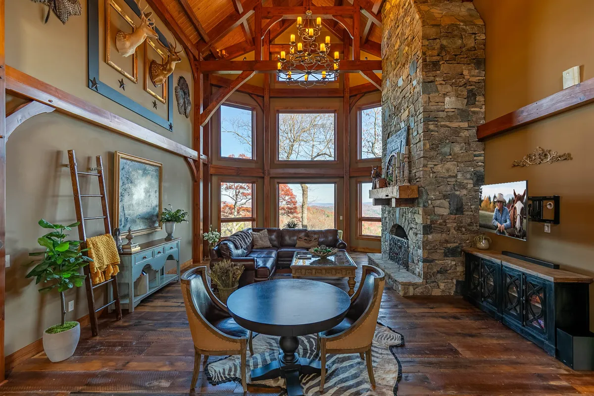 This image showcases a spacious great room with large windows that frame breathtaking mountain views. The stone fireplace becomes a focal point, surrounded by a comfortable leather sofa, rustic chandelier overhead, and decorative wall art. The ambiance is warm and inviting, perfect for family gatherings and cozy evenings.