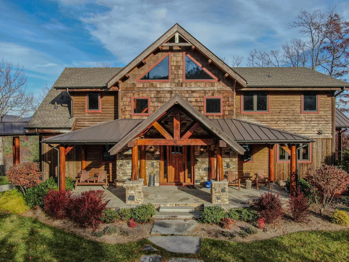 This image captures the property's exterior, featuring a welcoming wooden front door framed by sturdy stone columns. Large windows invite natural light inside, while a porch with comfortable seating overlooks the surrounding landscape. The scene hints at both the tranquility of the retreat and the potential for outdoor gatherings.