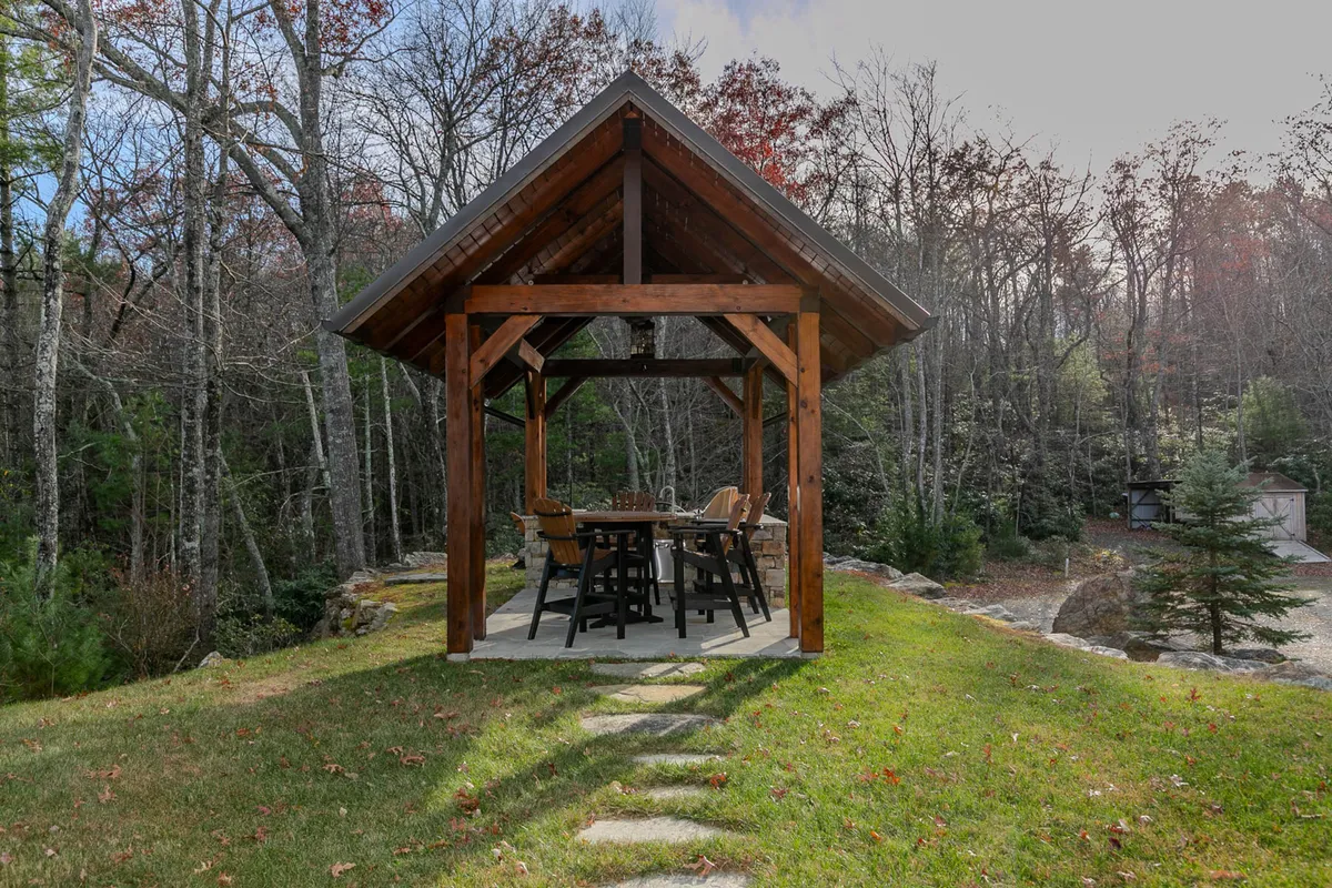 A peaceful outdoor scene featuring a wooden gazebo with a dining table set amidst mature trees. This inviting space is perfect for al fresco meals, relaxing in nature, and celebrating special occasions under the stars, embodying the lodge's promise of immersive seasonal experiences.