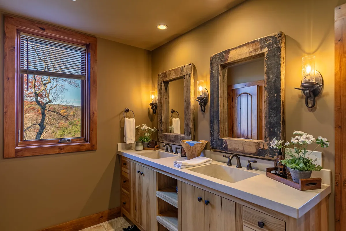 A serene, well-lit bathroom featuring a double sink vanity, rustic mirrors, warm wall sconces, and a window offering a soothing view. The calm, inviting mood emphasizes high-quality finishes and personal comfort.