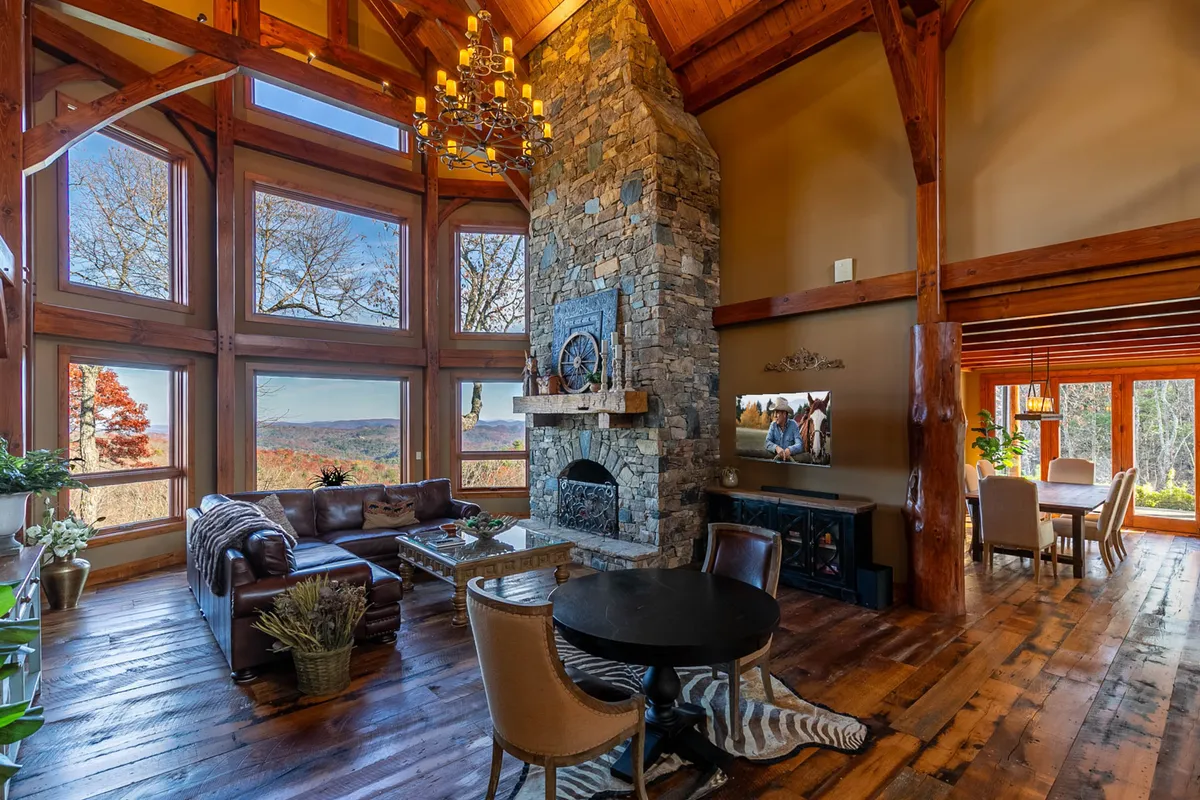 A beautifully appointed open-plan space featuring a large stone fireplace, expansive windows capturing the mountain vistas, a comfortable leather couch, and a rustic wooden dining table. The room is set for both intimate family time and festive gatherings.