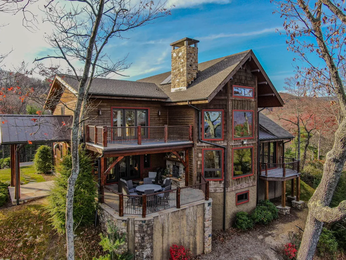 An exterior view of the lodge featuring a stone chimney, expansive windows, and a spacious deck area. The surrounding greenery and inviting deck highlight spaces perfect for relaxing after a day outdoors or gathering around the fire pit.