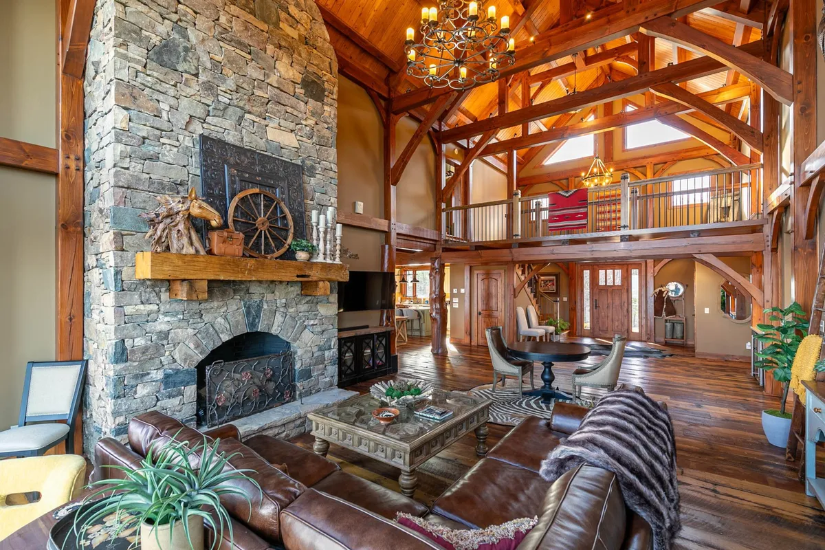 A cozy living room with a grand stone fireplace, exposed wooden beams, elegant chandelier, large windows letting in natural light, and comfortable seating. The scene conveys warmth, togetherness, and a welcoming atmosphere.