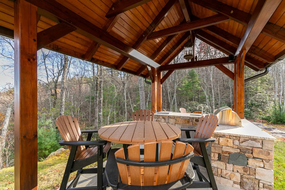 An outdoor kitchen setup sheltered by a wooden gazebo, featuring a circular wooden dining table and a stone countertop. Surrounded by lush greenery, this inviting area is perfect for barbecuing, socializing, and enjoying meals in nature.