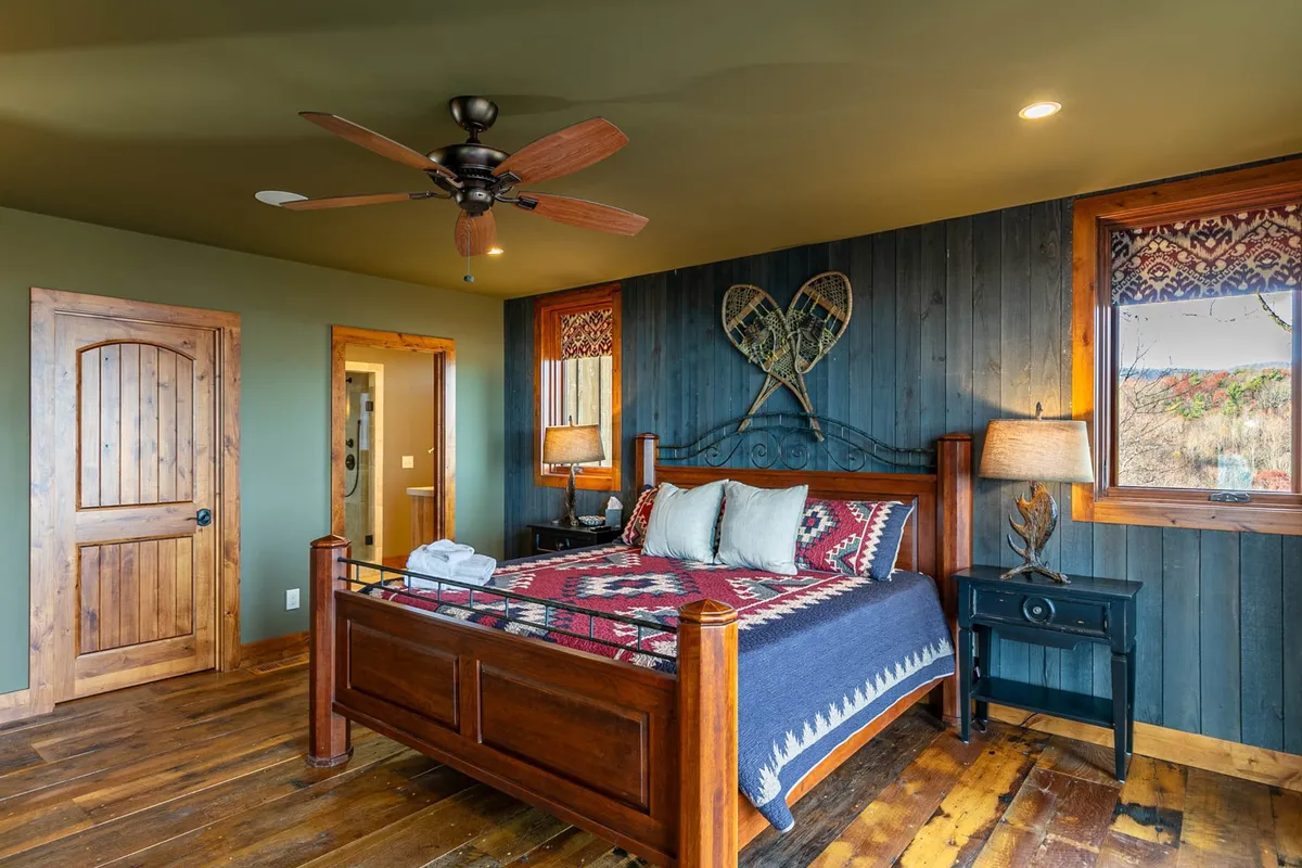A warmly lit bedroom with a wooden bed frame dressed in a colorful bedspread. Textured wall art and a nightstand with lamp lend a cozy, rustic vibe. Natural light streams through windows, creating a serene spot for rest and quiet moments.