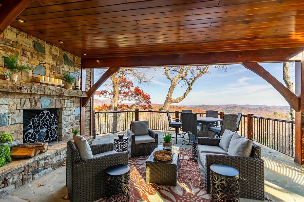 A beautiful exterior living space featuring a robust stone fireplace, inviting wicker seating area, and a dining table with chairs—all set against a breathtaking view of the mountains. The arrangement feels perfect for gathering, relaxing, and dining outdoors, with the natural landscape creating a tranquil backdrop.