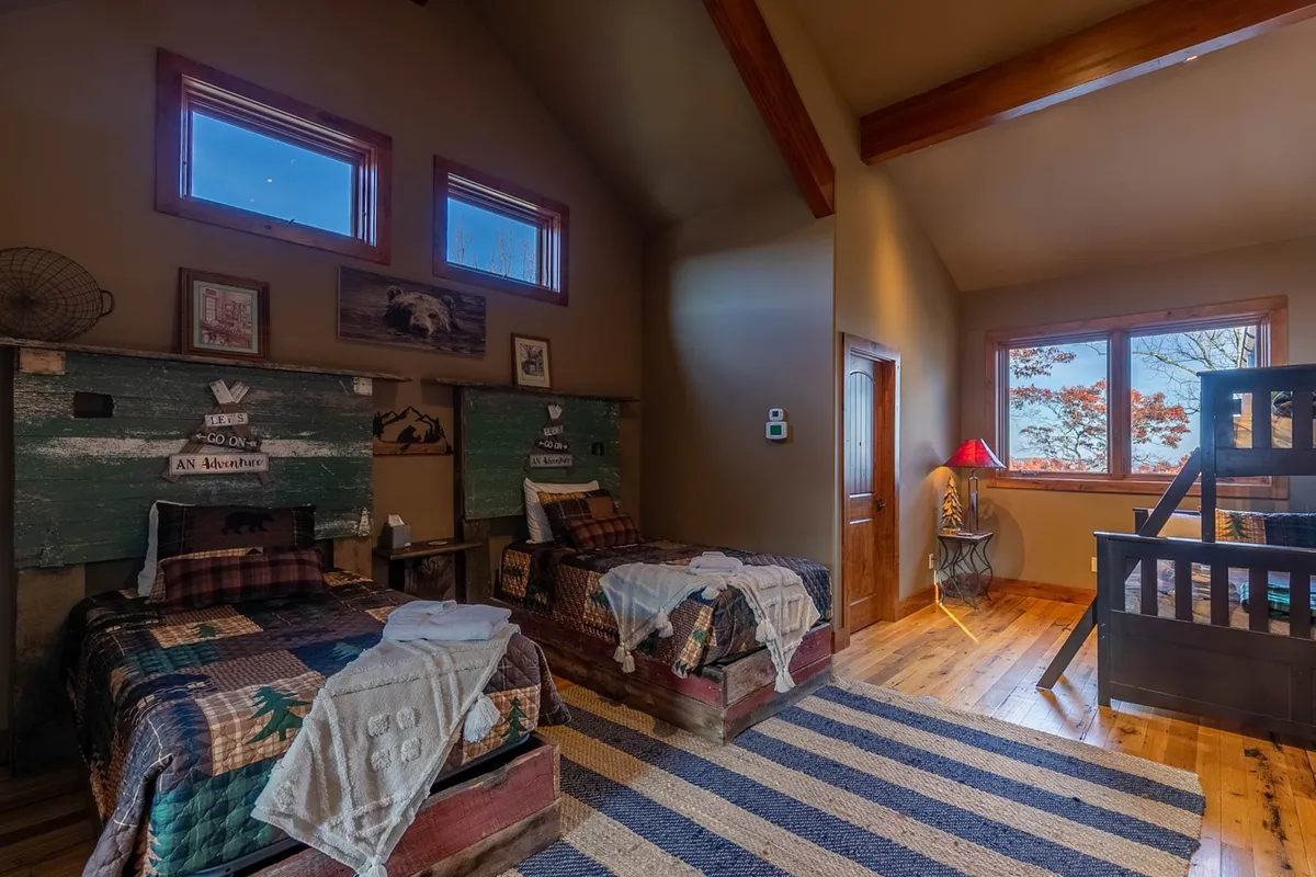 A vibrant bunkroom featuring wooden bunk beds and twin beds, all made up with inviting quilts. Wooden beams overhead and a large window allow sunlight to fill the space, making it ideal for relaxing, playing games, or sharing stories.